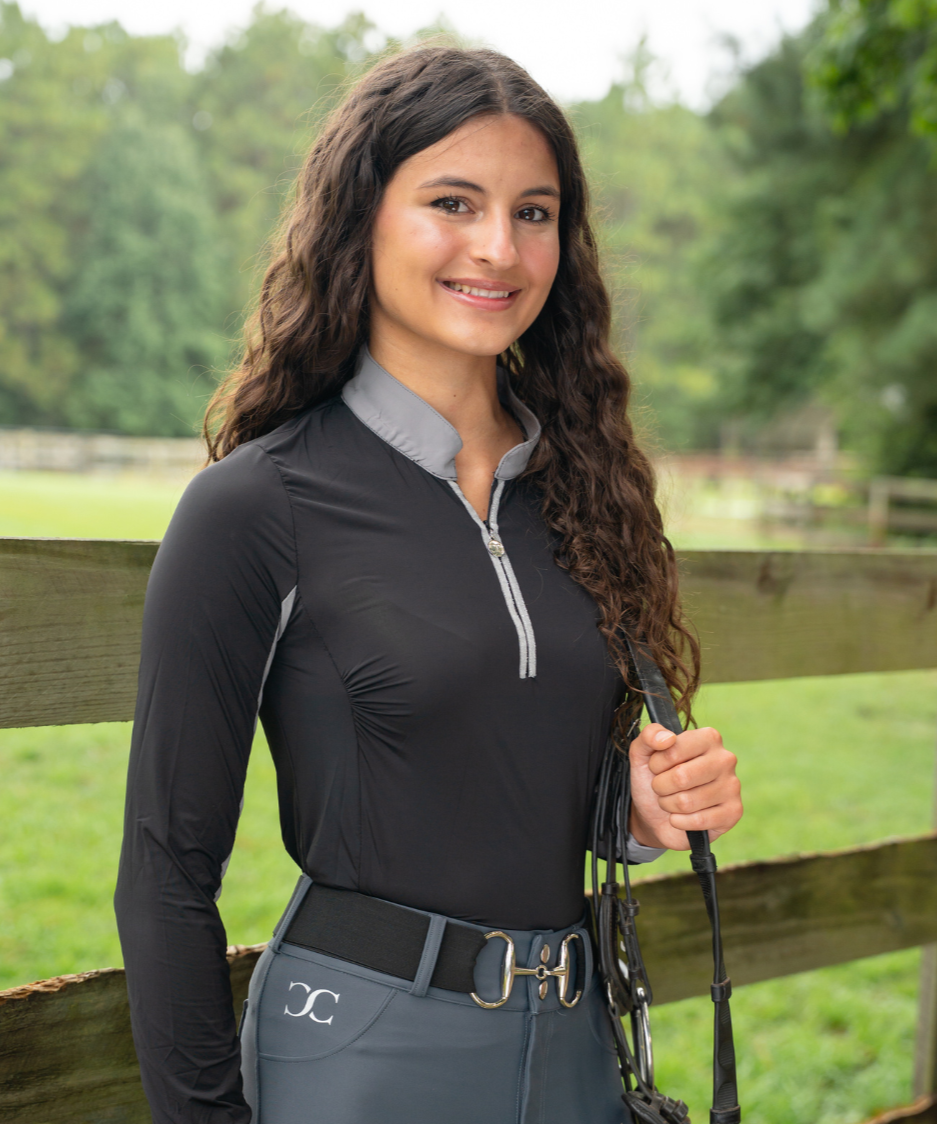 Woman in equestrian attire standing next to a wooden fence with a grassy field in the background.