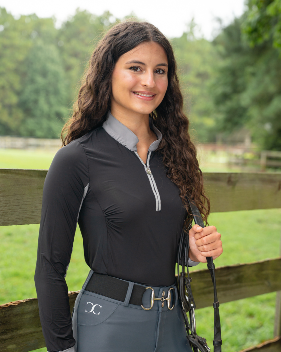 Woman in equestrian attire standing next to a wooden fence with a grassy field in the background.